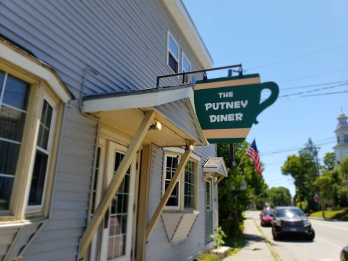Sign for "The Putney Diner" hanging from a building on a sunny day, with an American flag in the background.