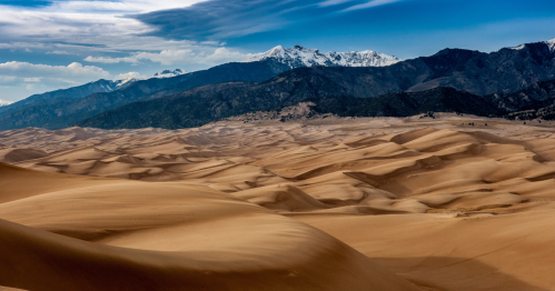 a sea of shadowy sand dunes leads the eye to snowcapped mountains in the background at great sand dunes national park in colorado