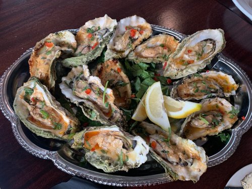 A silver platter filled with baked oysters topped with herbs, spices, and lemon wedges, garnished with greens.