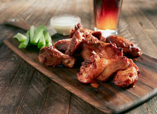 A wooden platter with glazed chicken wings, celery sticks, a small bowl of dipping sauce, and a drink in the background.