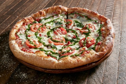 A freshly baked pizza topped with tomatoes, spinach, cheese, and herbs on a wooden serving board.