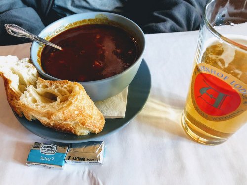 A bowl of soup with bread, butter packets, and a glass of beer on a table.