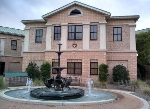 A brick building with large windows and a fountain featuring birds in front, surrounded by benches and greenery.