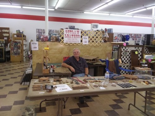 A man sits at a table displaying various leather products in a craft store. Signs indicate "Outlaw Leather."