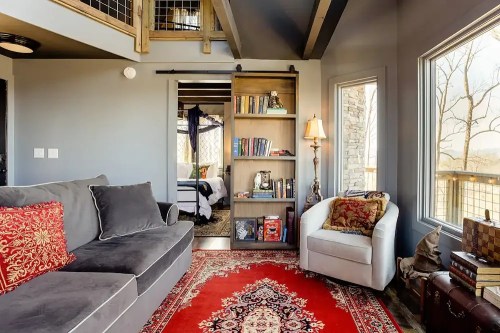 Cozy living space with a gray sofa, bookshelf, and armchair, featuring a red area rug and large windows.