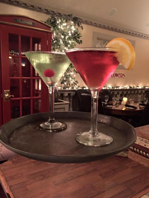 A tray holds two colorful cocktails: a green martini with a cherry and a red drink with a lemon slice.