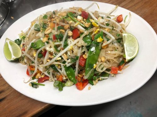 A plate of colorful pad thai with vegetables, peanuts, and lime wedges, garnished with fresh herbs.