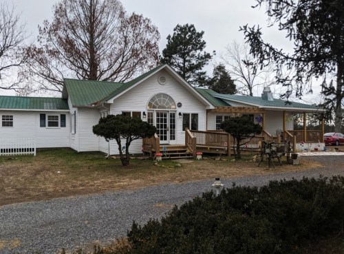 A white house with a green metal roof, surrounded by trees and shrubs, featuring a front porch and gravel driveway.