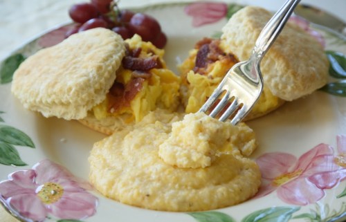A plate with two biscuit halves filled with eggs and bacon, alongside a serving of creamy grits and grapes.