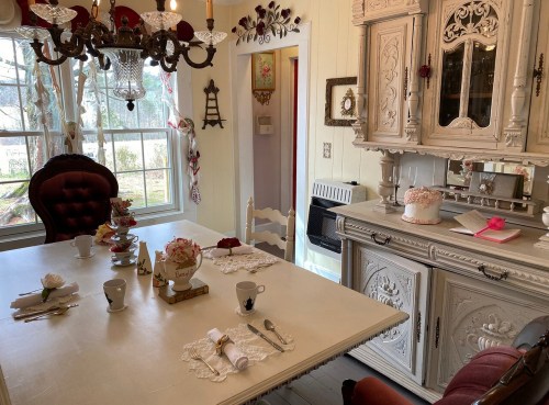 A cozy dining room with a vintage table set for tea, featuring floral decor and a charming chandelier.