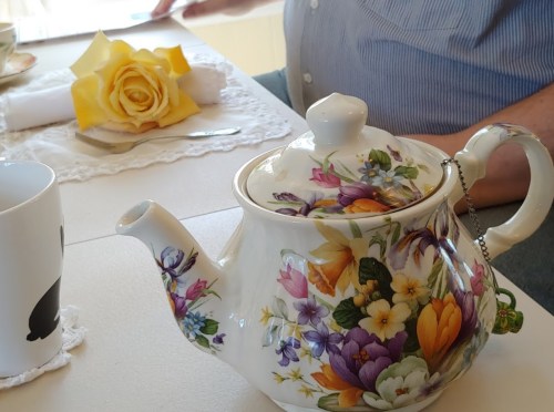 A floral teapot sits on a table beside a cup and a yellow rose, with a person’s hand visible in the background.