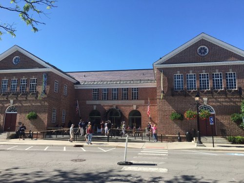 A brick building with large windows and flags, people gather outside on a sunny day.