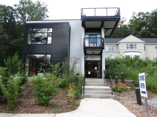 Modern building with large windows, a balcony, and a landscaped entrance, surrounded by greenery. Sign indicates "Store Open."