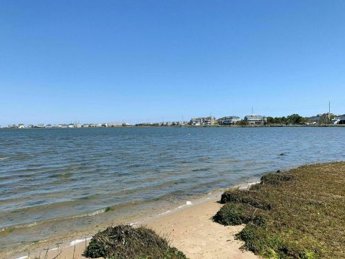 A calm shoreline with gentle waves, sandy beach, and distant houses under a clear blue sky.