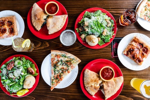 A spread of pizzas, calzones, salads, and drinks on red plates atop a wooden table.