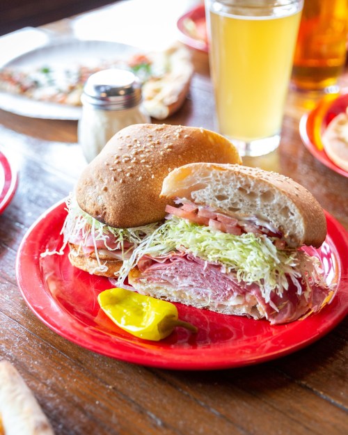 A close-up of a sandwich with layers of meat, lettuce, and tomato on a red plate, accompanied by a pickle and a drink.