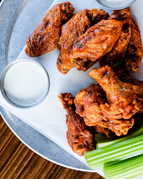 A platter of crispy chicken wings with two dipping sauces and celery sticks on a metal tray.