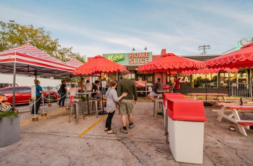 A vibrant outdoor pizza restaurant with red umbrellas, picnic tables, and customers enjoying the evening atmosphere.