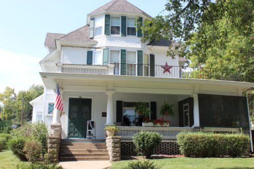 A charming two-story white house with a star decoration, porch, and American flag, surrounded by greenery.