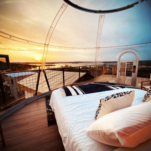 Cozy bedroom with a clear dome, offering a stunning sunset view over a lake and surrounding landscape.