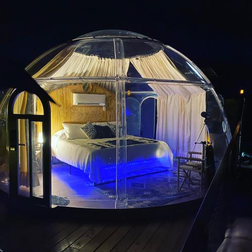 A cozy, illuminated transparent dome with a bed and soft curtains, set against a night sky.