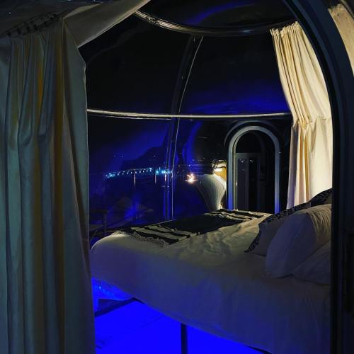 A cozy, modern bedroom with a glowing blue floor and sheer curtains, set in a glass structure with night views.