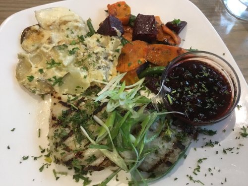 A plate featuring grilled fish, roasted vegetables, creamy potatoes, and a small bowl of sauce, garnished with herbs.