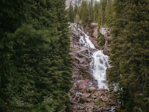 A cascading waterfall flows down rocky terrain, surrounded by lush green trees and a mountainous backdrop.