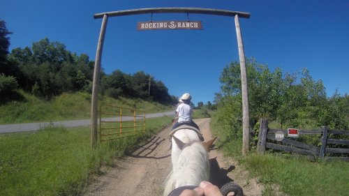 A person riding a horse approaches a sign that reads "Rocking S Ranch" on a sunny day. Green trees line the path.