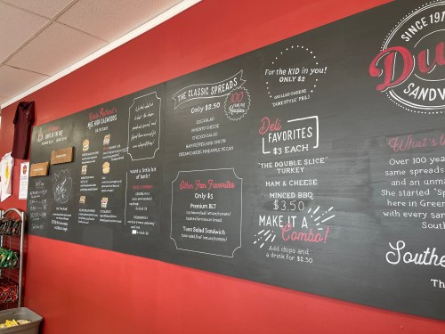 Menu board featuring sandwich options, prices, and specials in a casual dining setting. Red wall background.