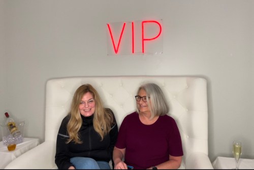 Two women sit on a white couch under a neon "VIP" sign, smiling and enjoying each other's company.