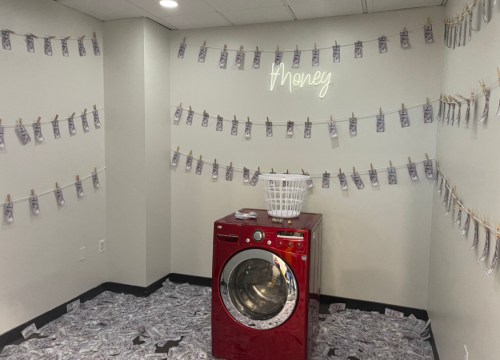 A laundry room with a red washing machine, decorated with fake money hanging on lines and scattered on the floor.