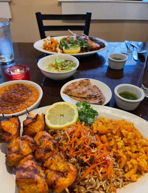 A table with two plates of colorful dishes, including grilled chicken, rice, salad, and desserts, with drinks in the background.