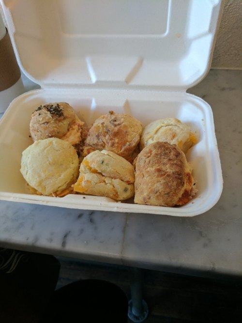 A takeout container filled with six assorted biscuits, some with cheese and herbs, on a marble surface.