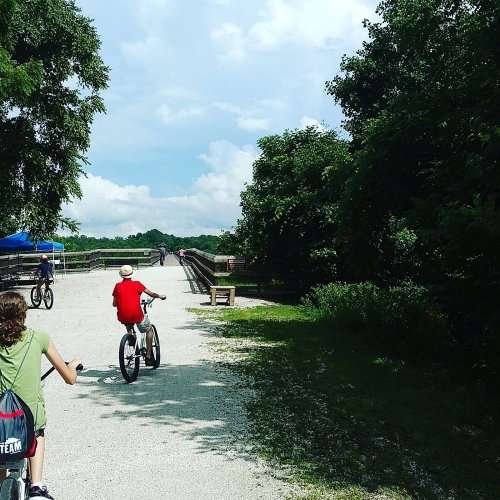 Two people ride bicycles along a path near a river, surrounded by trees and a blue sky with clouds.