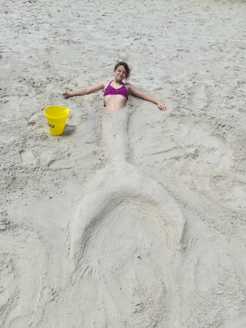A child in a bikini lies on the sand, with a sand mermaid tail sculpted around her. A yellow bucket is nearby.