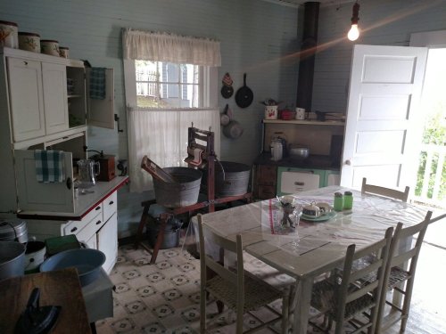A vintage kitchen with a wooden table, chairs, and various utensils, featuring a rustic charm and natural light.