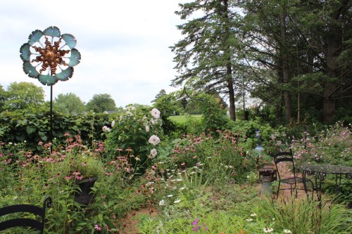 A lush garden filled with colorful flowers, a decorative wind sculpture, and a small seating area among greenery.