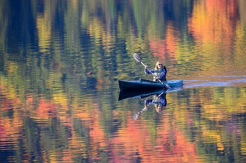 A person paddles a kayak on a calm lake, surrounded by vibrant autumn foliage reflected in the water.
