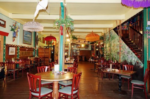 Cozy restaurant interior with wooden tables, colorful lamps, and tropical decor, featuring a staircase in the background.
