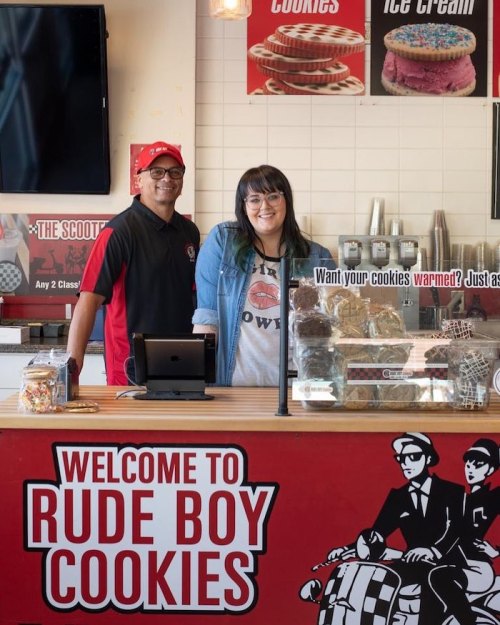 A smiling man and woman stand behind the counter of Rude Boy Cookies, with cookies and ice cream displayed.