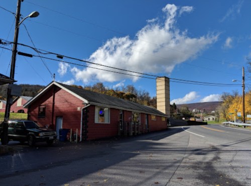 A red building with a tall chimney on a sunny day, surrounded by trees and mountains in the background.