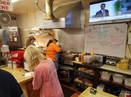 A busy kitchen with staff preparing food, a TV in the background, and a whiteboard displaying menu items and specials.