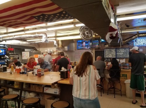 A busy diner with patrons at the counter, an American flag overhead, and a TV showing news in the background.