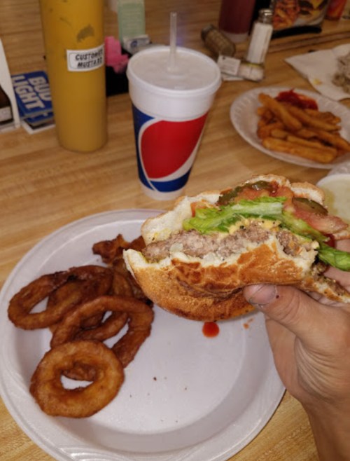 A hand holding a burger with lettuce and tomato, accompanied by onion rings and a soft drink on a plate.