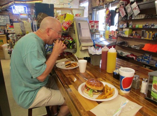 A man sits at a counter eating a burger and fries, with condiments and snacks visible in a small store.