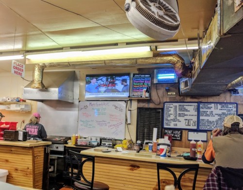 A cozy diner interior with a counter, menu board, TV, and handwritten notes on the wall.