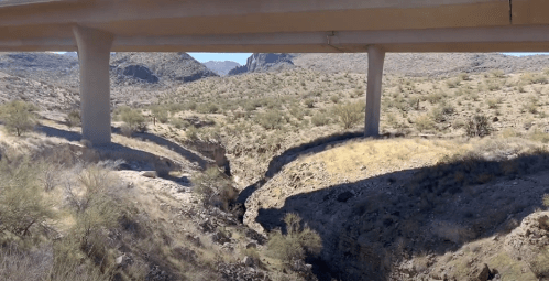 A view of a dry canyon beneath a highway overpass, surrounded by arid landscape and distant mountains.