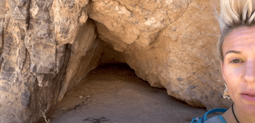 A rocky cave entrance with sandy ground, surrounded by large boulders and sparse vegetation.