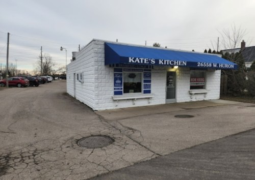 A small white building with a blue awning reading "Kate's Kitchen," located on a street with parked cars nearby.